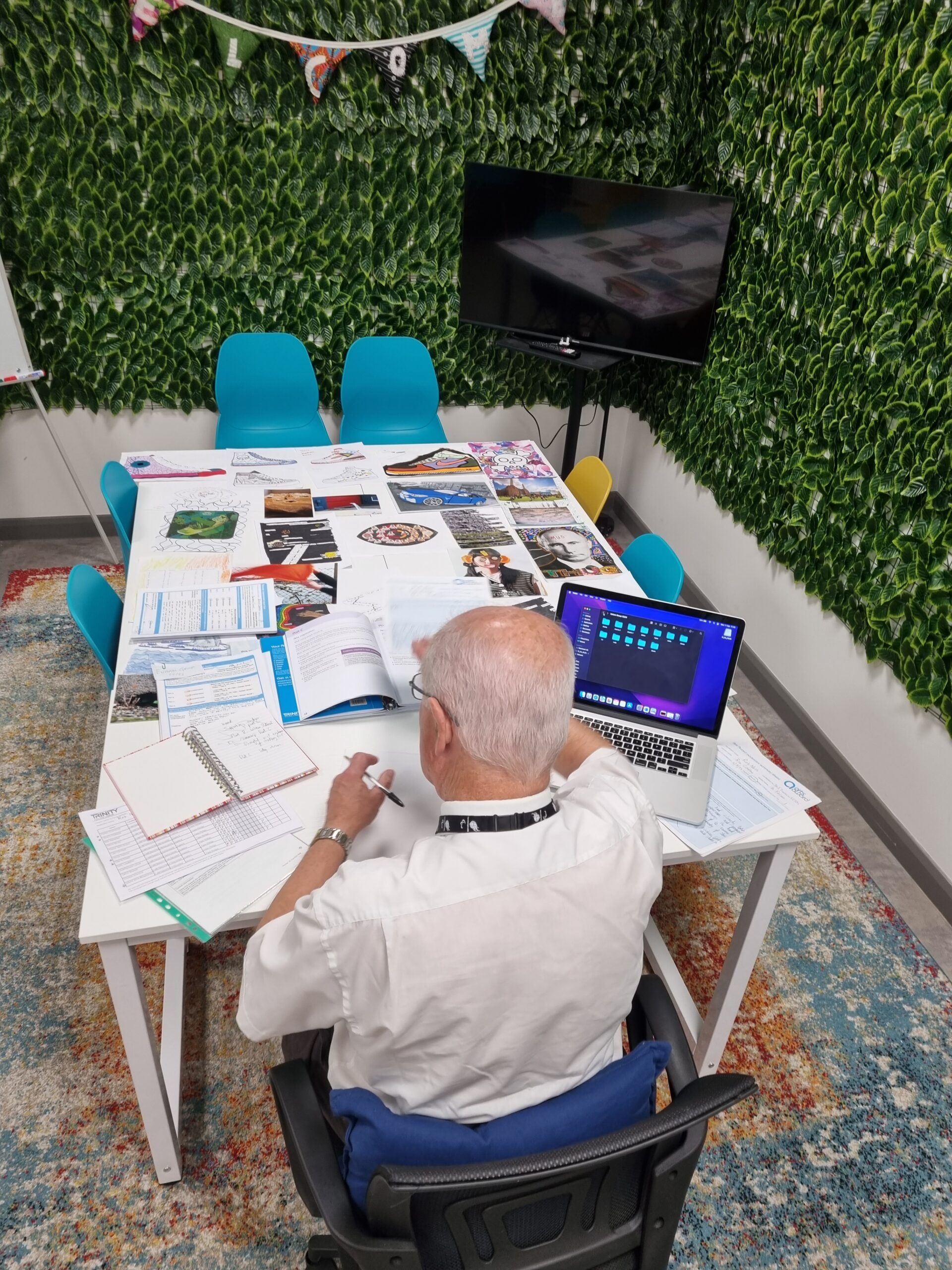 A man sits with his back tot he camera at a table. He is in the In Focus office space, marking a selection of artwork made by young people. The walls around him are covered in green leaves.
