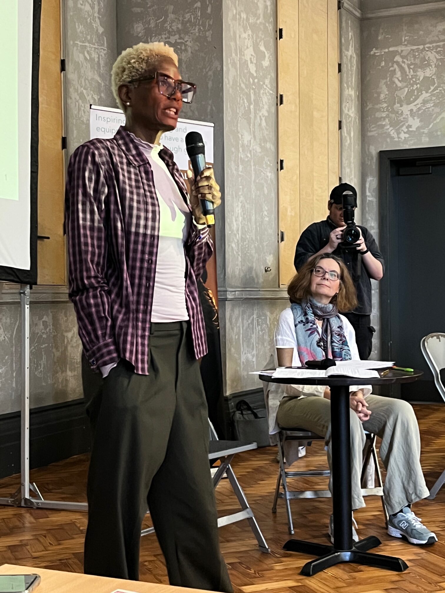 A black woman with short bleached hair and big black framed glasses stands in front of an assembly of people with a microphone. she is delivering a talk as the founder of Arts Inc in London at the Clore Leadership Now event.