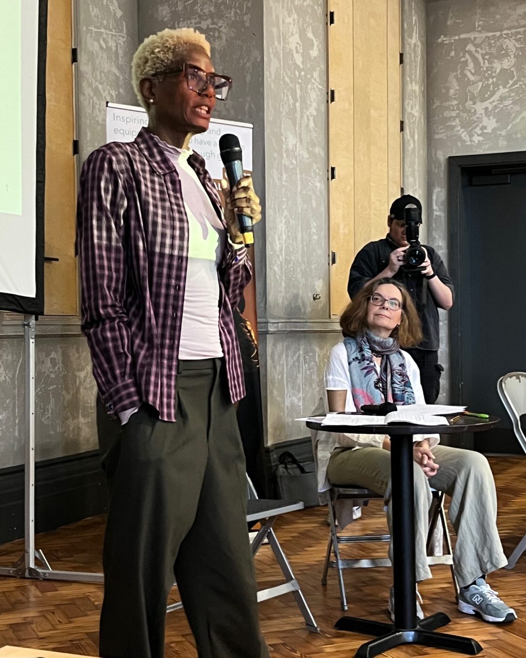 A black woman with short bleached hair and big black framed glasses stands in front of an assembly of people with a microphone. she is delivering a talk as the founder of Arts Inc in London at the Clore Leadership Now event.